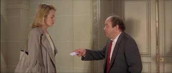 Movie still from “Le Dîner de Cons” (1998), directed by Francis Veber – A man in a business suit handing a piece of paper to a woman in a dress suit; Medium shot, Over the shoulder angle