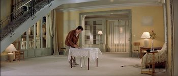 Movie still from “Le Dîner de Cons” (1998), directed by Francis Veber – A man standing in front of a table in a living room; Wide shot, High angle
