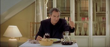 Movie still from “Le Dîner de Cons” (1998), directed by Francis Veber – A man sitting at a table with a plate of food and a glass of wine in front of him; Medium shot, Low angle