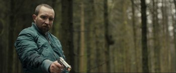 Movie still from “The Disappearance of Alice Creed” (2009), directed by J Blakeson – A man holding a gun while standing next to a forest; Medium shot, Over the shoulder angle