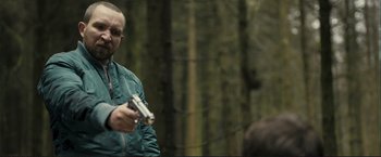 Movie still from “The Disappearance of Alice Creed” (2009), directed by J Blakeson – A man holding a gun while standing next to a forest; Medium shot, Over the shoulder angle