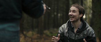 Movie still from “The Disappearance of Alice Creed” (2009), directed by J Blakeson – A man in the woods holding a gun and laughing; Medium shot, Over the shoulder angle