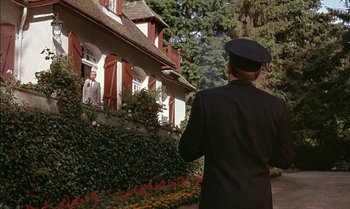 Movie still from “The Discreet Charm of the Bourgeoisie” (1972), directed by Luis Buñuel – A man standing in front of a house with flowers around it; Wide shot, Low angle