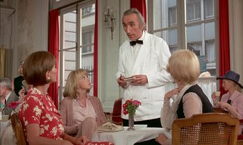 Movie still from “The Discreet Charm of the Bourgeoisie” (1972), directed by Luis Buñuel – A man in a white jacket is talking to a group of people; Medium shot, Over the shoulder angle