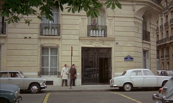 Movie still from “The Discreet Charm of the Bourgeoisie” (1972), directed by Luis Buñuel – Two men standing on the sidewalk in front of a building; Extreme Wide shot, High angle