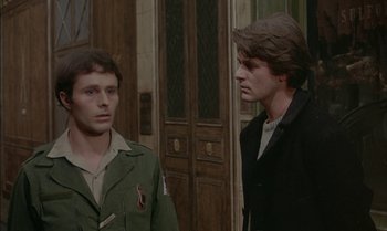 Movie still from “The Discreet Charm of the Bourgeoisie” (1972), directed by Luis Buñuel – Two men standing next to each other in front of a building; Medium shot, Over the shoulder angle