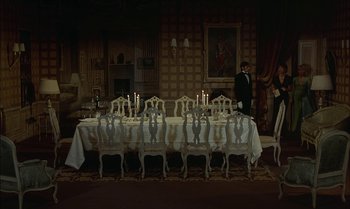Movie still from “The Discreet Charm of the Bourgeoisie” (1972), directed by Luis Buñuel – A man standing in front of a long table; Wide shot, High angle