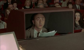 Movie still from “The Discreet Charm of the Bourgeoisie” (1972), directed by Luis Buñuel – A man sitting in front of an audience holding papers; Medium shot, High angle