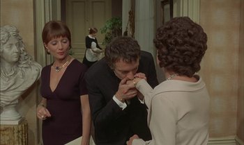 Movie still from “The Discreet Charm of the Bourgeoisie” (1972), directed by Luis Buñuel – A man and a woman standing next to each other in front of another woman; Medium shot, Over the shoulder angle
