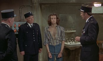 Movie still from “The Discreet Charm of the Bourgeoisie” (1972), directed by Luis Buñuel – A man in a striped shirt is being handcuffed by two police officers; Medium shot, High angle