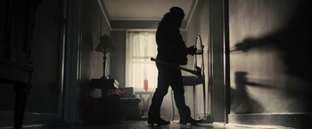 Movie still from “The Domestics” (2018), directed by Mike P. Nelson – A person holding a hammer in a living room; Wide shot, Low angle