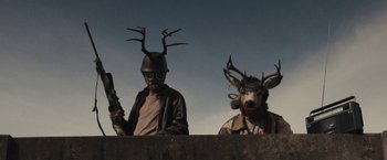 Movie still from “The Domestics” (2018), directed by Mike P. Nelson – A man with a gun and a man with a deer head on his head; Medium shot, Over the shoulder angle