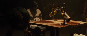 Movie still from “The Domestics” (2018), directed by Mike P. Nelson – A man sitting at a wooden table with a knife in his hand; Medium shot, Over the shoulder angle