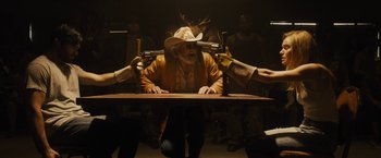 Movie still from “The Domestics” (2018), directed by Mike P. Nelson – A group of people sitting at a table holding guns; Medium shot, Over the shoulder angle