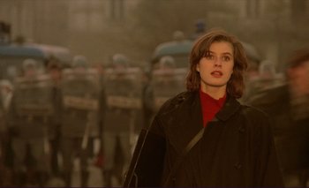 A woman in a red shirt and black jacket standing in front of a line of police