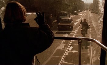 Movie still from “The Double Life of Véronique” (1991), directed by Krzysztof Kieslowski – A person is taking a picture of a street with cars; Wide shot, Over the shoulder angle