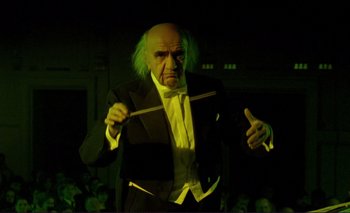 Movie still from “The Double Life of Véronique” (1991), directed by Krzysztof Kieslowski – An old man dressed in a suit and wig holding a conductor's baton; Medium shot, Low angle