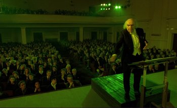 Movie still from “The Double Life of Véronique” (1991), directed by Krzysztof Kieslowski – A man in a suit and wig standing on a stage in front of an auditorium full of people; Wide shot, Low angle