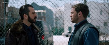Movie still from “The Drop” (2014), directed by Michaël R. Roskam – Two men standing next to each other in front of a chain link fence; Medium shot, Over the shoulder angle