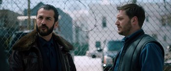 Movie still from “The Drop” (2014), directed by Michaël R. Roskam – Two men standing next to each other in front of a chain link fence; Close Up shot, Over the shoulder angle