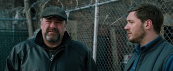 Movie still from “The Drop” (2014), directed by Michaël R. Roskam – A man wearing a hat standing in front of a fence; Close Up shot, Over the shoulder angle
