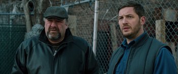 Movie still from “The Drop” (2014), directed by Michaël R. Roskam – Two men standing next to each other in front of a chain link fence; Close Up shot, Over the shoulder angle