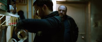 Movie still from “The Drop” (2014), directed by Michaël R. Roskam – A man standing next to another man in a dark room; Medium shot, Over the shoulder angle