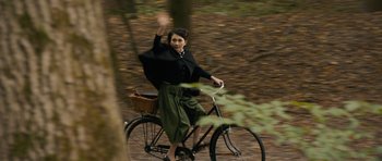 Movie still from “The Duke of Burgundy” (2014), directed by Peter Strickland – A woman riding a bike through the woods waving; Wide shot, High angle