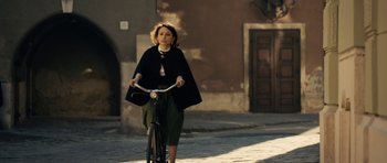 Movie still from “The Duke of Burgundy” (2014), directed by Peter Strickland – A woman riding a bike down a street; Wide shot, Over the shoulder angle
