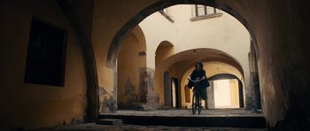 Movie still from “The Duke of Burgundy” (2014), directed by Peter Strickland – A person riding a bike through an archway; Extreme Wide shot, Low angle
