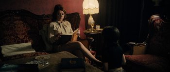 Movie still from “The Duke of Burgundy” (2014), directed by Peter Strickland – A woman sitting in a chair reading a book to a young girl; Medium shot, Over the shoulder angle