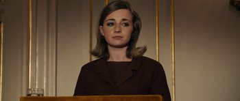 Movie still from “The Duke of Burgundy” (2014), directed by Peter Strickland – A woman in a brown jacket is standing in front of a podium; Close Up shot, Low angle