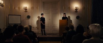 Movie still from “The Duke of Burgundy” (2014), directed by Peter Strickland – Two women standing in front of a podium in front of a crowd of onlookers; Wide shot, Over the shoulder angle