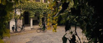 Movie still from “The Duke of Burgundy” (2014), directed by Peter Strickland – A person riding a bike on a paved street; Extreme Wide shot, High angle