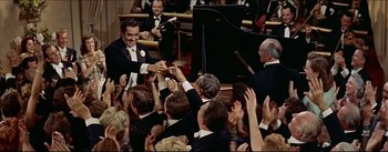 Movie still from “The Eddy Duchin Story” (1956), directed by George Sidney – A group of people in formal wear holding hands and clapping in front of an audience; Wide shot, High angle