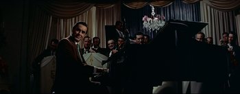 Movie still from “The Eddy Duchin Story” (1956), directed by George Sidney – A group of men standing around a grand piano; Medium shot, Over the shoulder angle