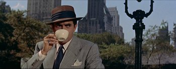 Movie still from “The Eddy Duchin Story” (1956), directed by George Sidney – A man wearing a suit and hat drinking from a white cup; Medium shot, Over the shoulder angle