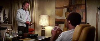 Movie still from “The Enforcer” (1976), directed by James Fargo – A man sitting in front of a table holding a wine glass; Medium shot, Over the shoulder angle