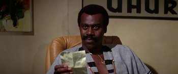 Movie still from “The Enforcer” (1976), directed by James Fargo – A man sitting in a chair holding a piece of paper; Close Up shot, Low angle