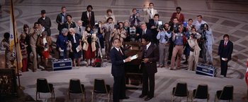 Movie still from “The Enforcer” (1976), directed by James Fargo – Two men in business suits holding papers in front of an audience; Wide shot, High angle