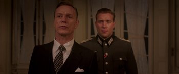 Movie still from “The Exception” (2016), directed by David Leveaux – A man in a suit standing next to another man in a uniform; Medium shot, Low angle
