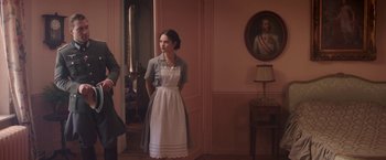 Movie still from “The Exception” (2016), directed by David Leveaux – A woman standing in front of a wall with a painting on the wall; Medium shot, Over the shoulder angle