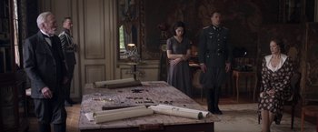 Movie still from “The Exception” (2016), directed by David Leveaux – A man and a woman standing in front of a table; Wide shot, High angle