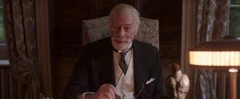 Movie still from “The Exception” (2016), directed by David Leveaux – An older man sitting in a chair wearing a suit and tie; Close Up shot, Low angle