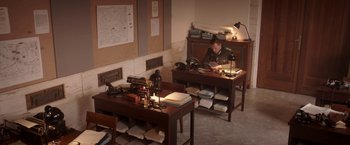 Movie still from “The Exception” (2016), directed by David Leveaux – A man sitting at a desk in front of an old fashioned sewing machine; Wide shot, High angle