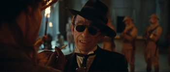 Movie still from “The Extraordinary Adventures of Adèle Blanc-Sec” (2010), directed by Luc Besson – An older man wearing a hat and sunglasses smoking a cigarette; Close Up shot, Low angle