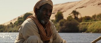 Movie still from “The Extraordinary Adventures of Adèle Blanc-Sec” (2010), directed by Luc Besson – An older man wearing a turban sitting in front of a body of water; Medium shot, Over the shoulder angle