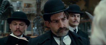 Movie still from “The Extraordinary Adventures of Adèle Blanc-Sec” (2010), directed by Luc Besson – A man with a mustache wearing a hat and a tie; Close Up shot, Over the shoulder angle