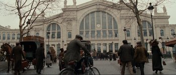 Movie still from “The Extraordinary Adventures of Adèle Blanc-Sec” (2010), directed by Luc Besson – A man riding a bike in front of a large building; Extreme Wide shot, Low angle
