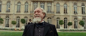 Movie still from “The Extraordinary Adventures of Adèle Blanc-Sec” (2010), directed by Luc Besson – An old man with a beard and a suit and bow tie in front of an old building; Medium shot, Low angle